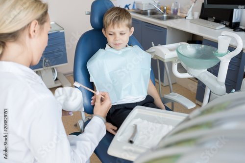 Portrait of brave little boy sitting in dental chair and looking at female dentist explaining how to brush teeth  copy space