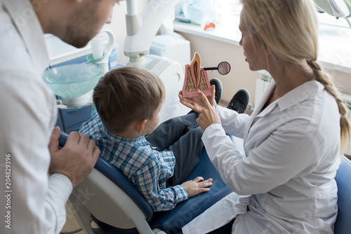Portrait of female dentist holding tooth model showing it to little boy sitting in dental chair with father at his side  copy space