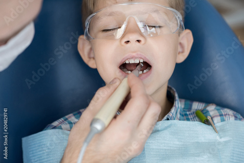 Close up portrait of cute little boy sitting in dental chair with mouth open while dentist treating his teeth and filling cavity in modern clinic
