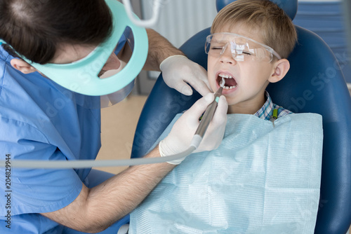 Portrait of cute little boy sitting in dental chair and wincing with pain while dentist treating his teeth and filling cavity in modern clinic