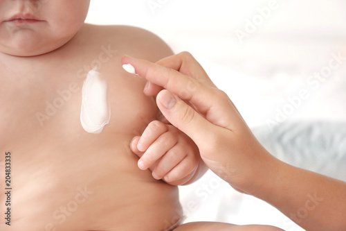 Woman applying body cream on her baby against light background