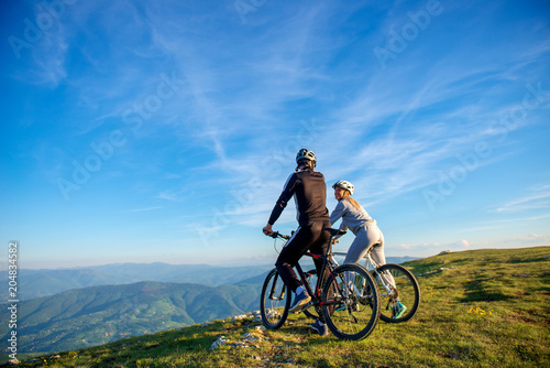Cyclist couple with mountain bikes standing on the hill under the evening sky and enjoying bright sun at the sunset. © FS-Stock
