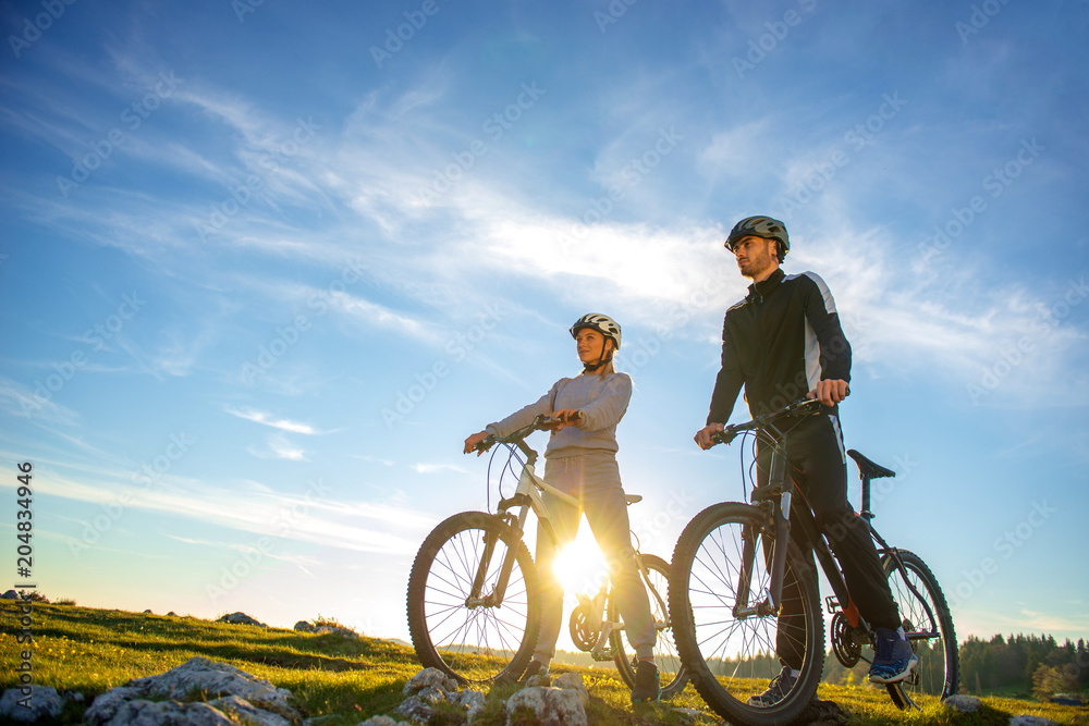 Cyclist couple with mountain bikes standing on the hill under the evening sky and enjoying bright sun at the sunset. Cyclist couple with mountain bikes standing on the hill under the evening sky and enjoying bright sun at the sunset.