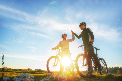 Fototapeta Naklejka Na Ścianę i Meble -  happy couple goes on a mountain road in the woods on bikes with helmets giving each other a high five