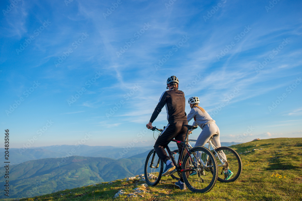 Obraz premium Cyclist couple with mountain bikes standing on the hill under the evening sky and enjoying bright sun at the sunset.