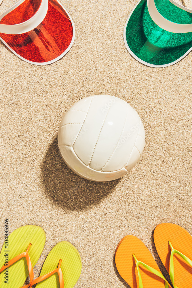 flat lay with white volleyball ball, colorful caps and flip flops arranged on sand