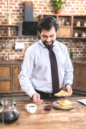 smiling handsome loner businessman adding jam on toast at kitchen
