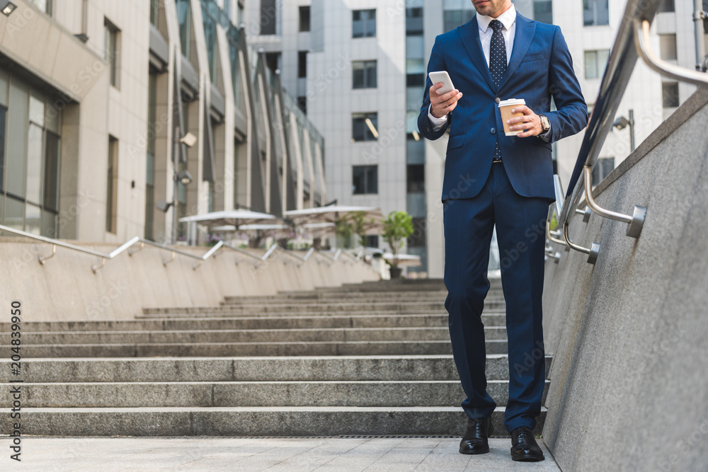 cropped shot of businessman in stylish suit with coffee to go using smartphone while going down stairs