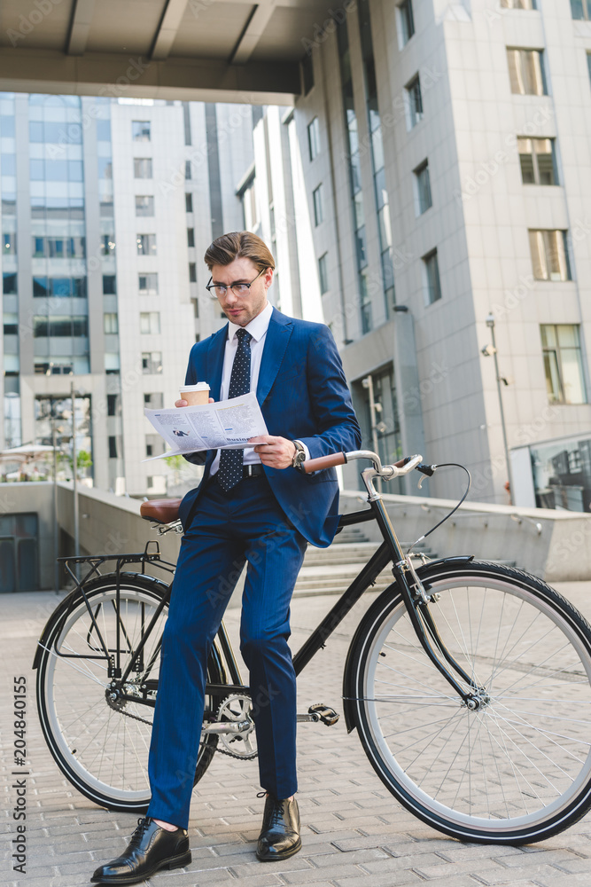 businessman in stylish suit with newspaper and paper cup of coffee leaning on bicycle businessman in stylish suit with newspaper and paper cup of coffee leaning on bicycle