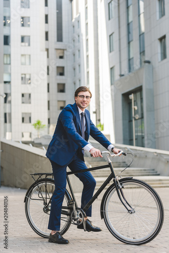 Fototapeta Naklejka Na Ścianę i Meble -  handsome young businessman in stylish suit on bicycle in business district