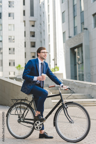 handsome young businessman with paper cup of coffee on bicycle in business district