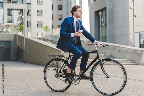 young businessman in stylish suit with coffee to go sitting on vintage bicycle