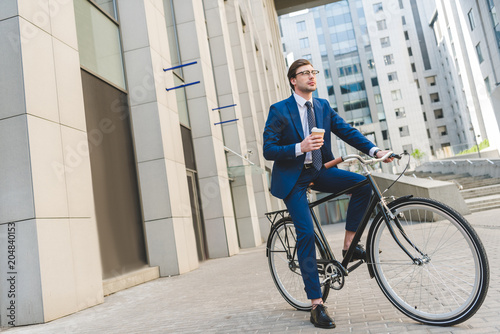 handsome young businessman in stylish suit with coffee to go sitting on vintage bicycle