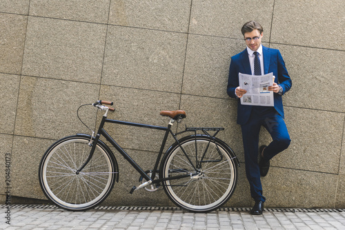 handsome young businessman in stylish suit with bicycle reading newspaper leaning on wall