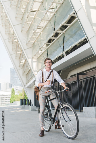 attractive young man with paper cup of coffee on vintage bicycle near sports stadium building