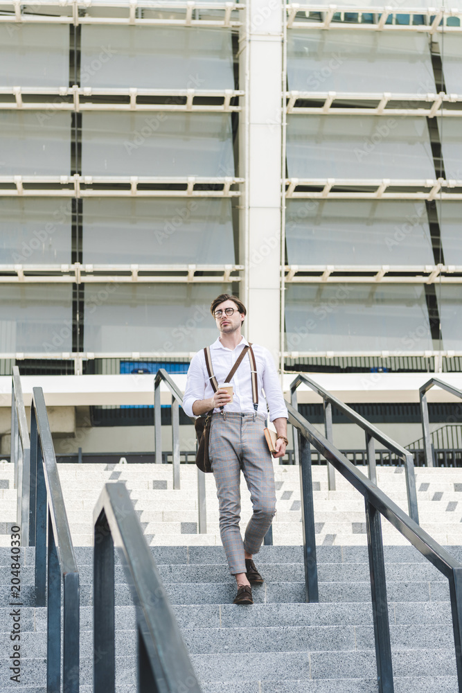 stylish young man walking down stairs in front of stadium with book and coffee to go