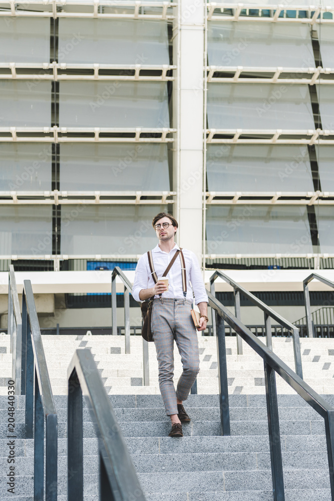 stylish young man walking down stairs in front of stadium with book and coffee to go stylish young man walking down stairs in front of stadium with book and coffee to go
