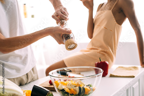 cropped shot of man using pepper grinder over bowl with salad and woman sitting on table at kitchen
