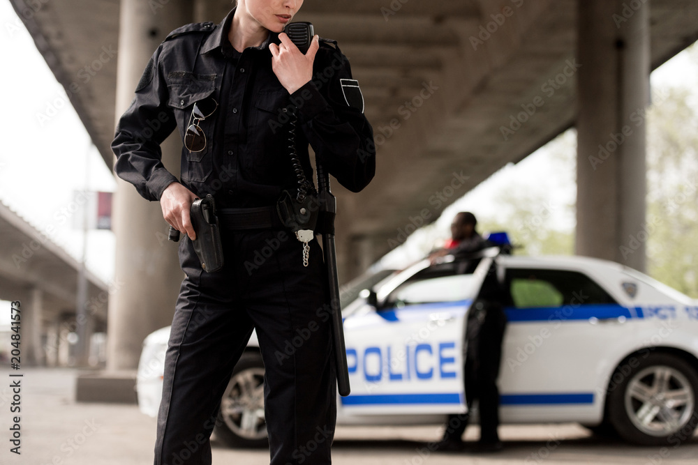 cropped shot of policewoman using walkie-talkie with blurred partner near car on background cropped shot of policewoman using walkie-talkie with blurred partner near car on background