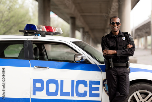handsome african american police officer with crossed arms leaning back on car and looking at camera