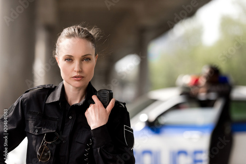 Canvas Print policewoman using walkie-talkie and looking at camera with blurred partner near