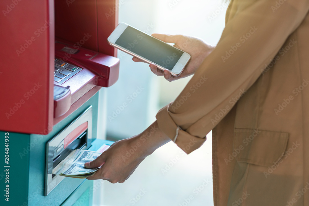 Naklejka premium woman using the smart mobile phone for withdrawing in front of the ATM, business Automatic Teller Machine concept