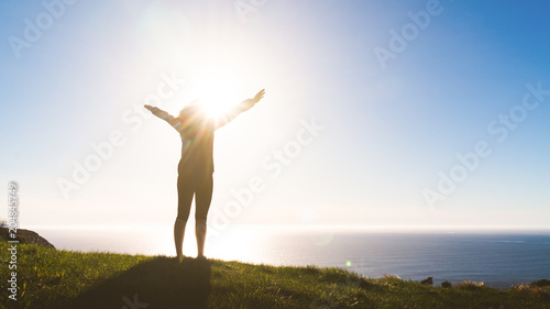 Excited woman stand on mountain hill with hands raised looking towards the ocean. Global female empowerment. Travel highlight in nature.