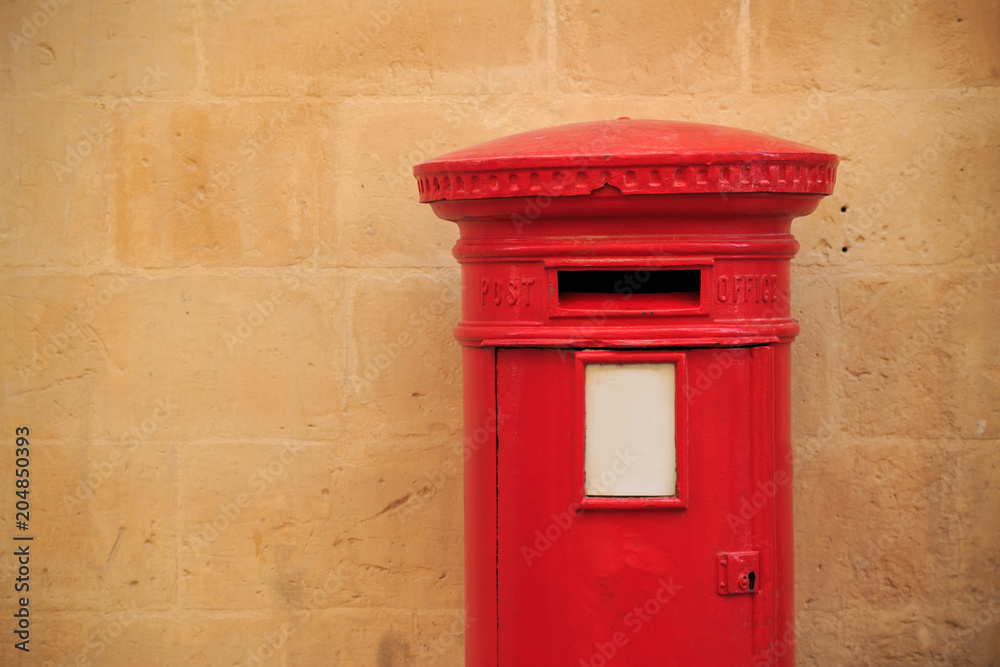 Traditional, British postbox in red color. Mailbox that receives the letters. Light orange limestone wall for background. Copyspace, close up view.