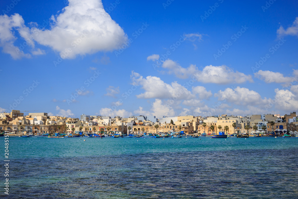 Marsaxlokk historic port full of boats in Malta. Blue sky with few white clouds and village background. Panoramic view.