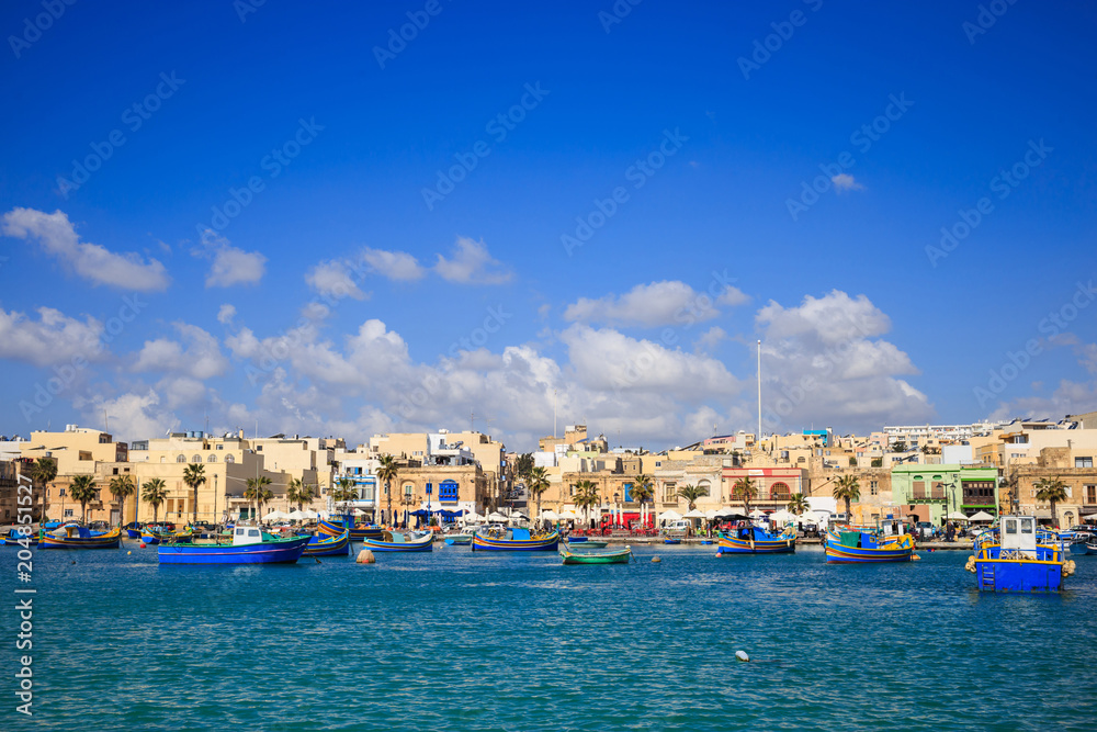 Marsaxlokk historic port full of boats in Malta. Blue sky with few white clouds and village background. Panoramic view.