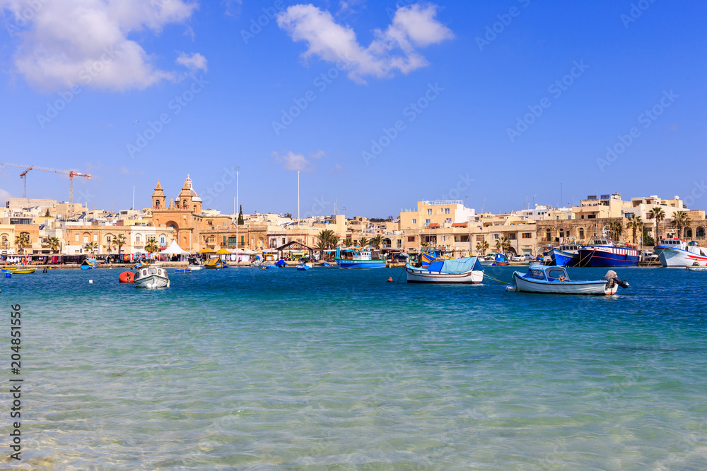 Marsaxlokk historic port with many boats in transparent sea, Malta. Blue sky and village background.