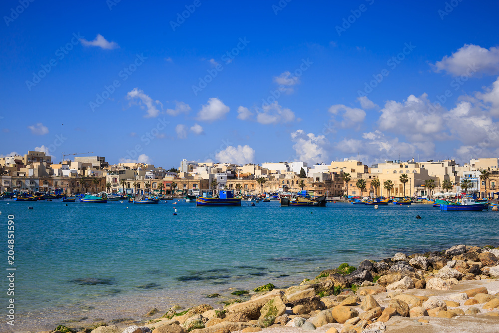 Marsaxlokk historic port with many boats in transparent sea and beach with stones, Malta. Blue sky and village background.