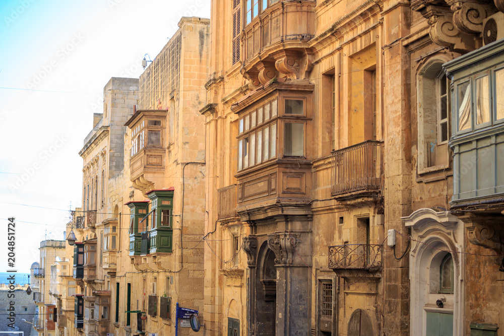 Malta, Valletta, traditional sandstone buildings with colorful wooden windows on balconies. Blue sky with clouds and sea background.