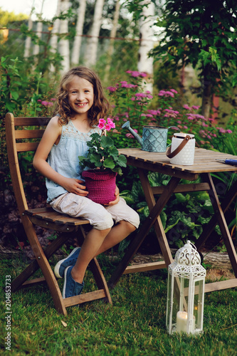 happy kid girl playing in summer garden, holding heranium flower in pot. Teaching kids to love nature and take care about plants photo