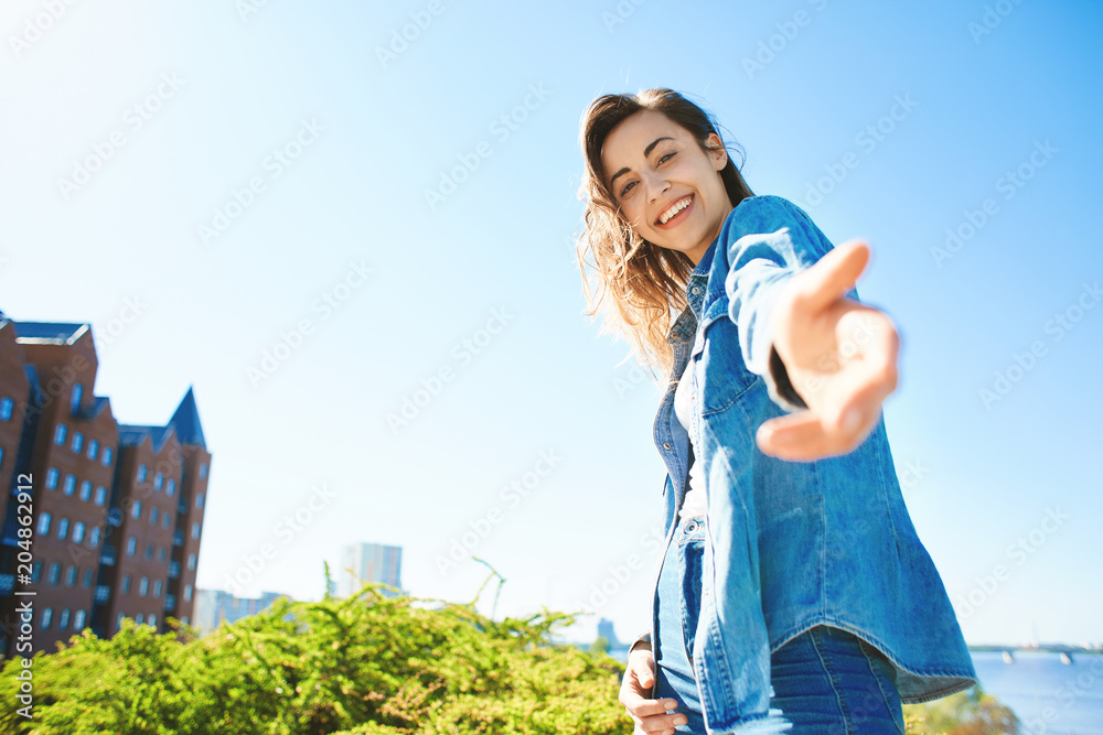 Obraz premium portrait of a young smiling attractive woman in jeans clothes at sunny day on the blue sky background. happy woman gives a hand to someone like follow me . first person view.