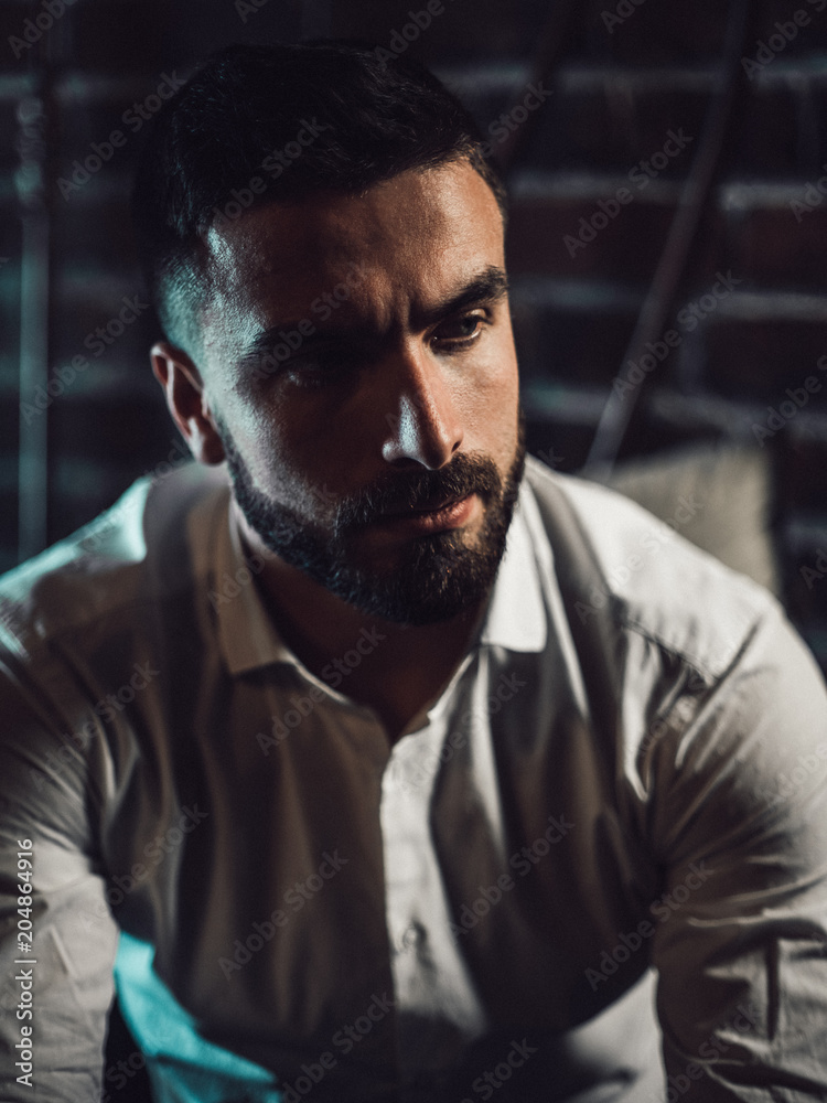 A handsome man with a beard and a white shirt sits on the bed and looks thoughtfully at the camera. Intimate atmosphere and twilight. A handsome man with a beard and a white shirt sits on the bed and looks thoughtfully at the camera. Intimate atmosphere and twilight.