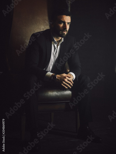 A handsome man with a beard and in a classic suit sits in a leather chair and looks at the camera. 