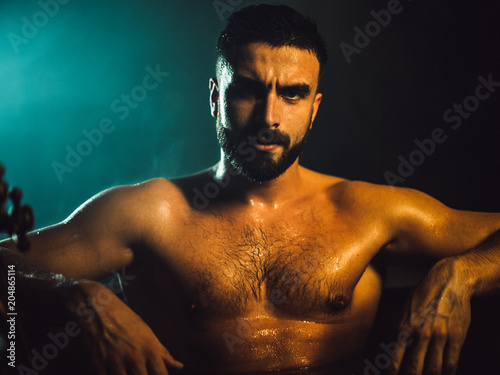 A handsome, muscular man with a beard lies in a bronze bathroom. Steam coming from the hot water. He looks at the camera, drops of water running down the body