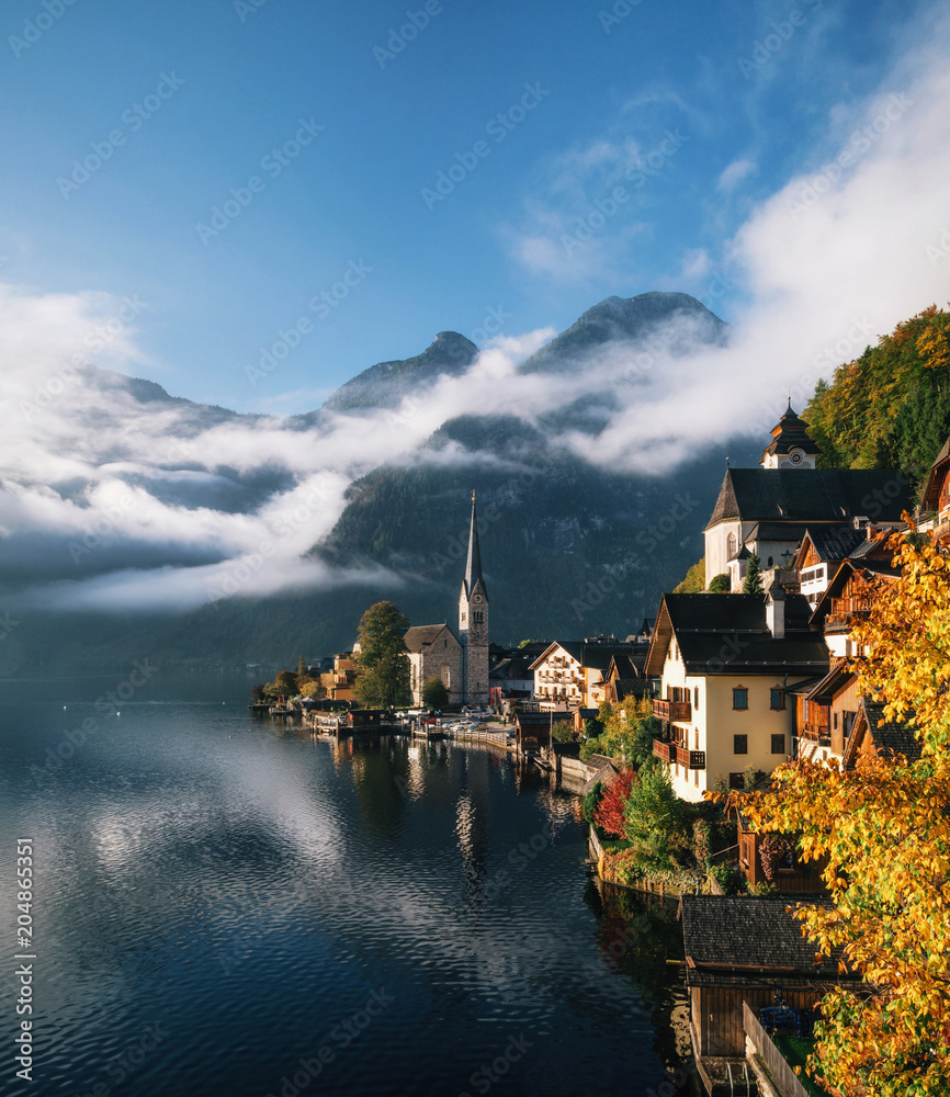 Fototapeta premium Malowniczy widok na słynne miasto nad jeziorem Hallstatt odbijające się w jeziorze Hallstattersee w austriackich Alpach w porannym świetle z jasnymi chmurami, region Salzkammergut, Austria