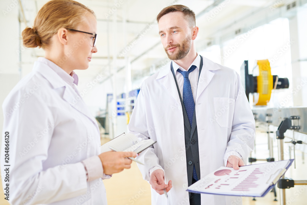 Bearded middle-aged dairy plant worker and his pretty colleague wearing white coats standing at modern production department while taking inventory Bearded middle-aged dairy plant worker and his pretty colleague wearing white coats standing at modern production department while taking inventory