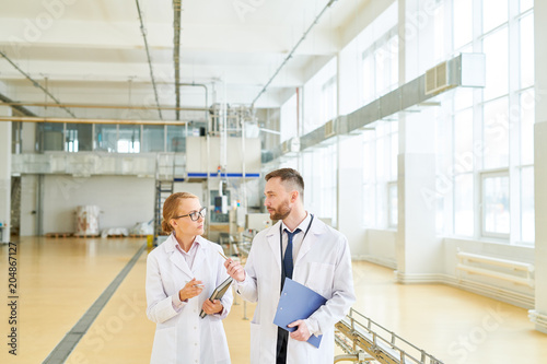 Bearded middle-aged technologist wearing white coat explaining details of production process to female inspector while walking along spacious dairy factory