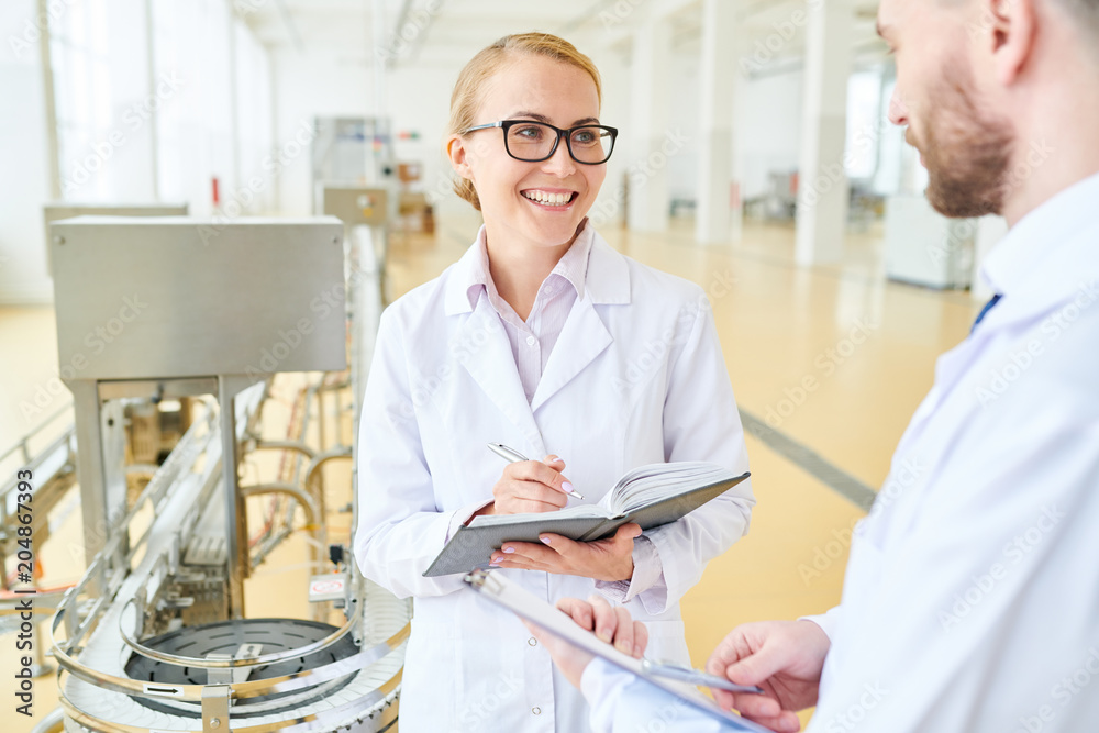 Obraz premium Portrait shot of attractive technologist with charming smile talking to male colleague and taking notes while having working meeting at spacious production department of milk factory.