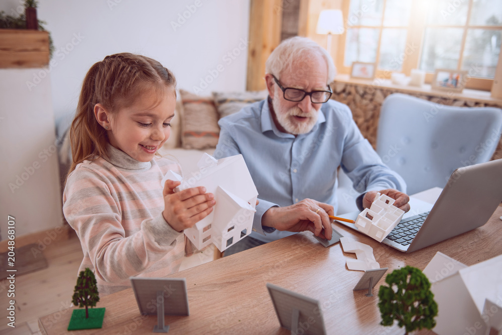 So interesting. Nice cheerful girl holding a house model while standing near her grandfather So interesting. Nice cheerful girl holding a house model while standing near her grandfather