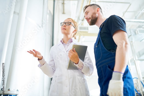 Pretty fair-haired factory worker wearing white coat discussing equipment occurrences with bearded technician while standing at spacious production department  low angle view