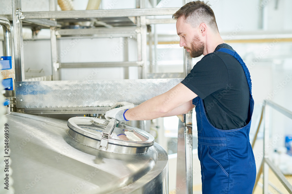 Profile view of bearded dairy factory worker wearing overall standing at production department and controlling technological process Profile view of bearded dairy factory worker wearing overall standing at production department and controlling technological process