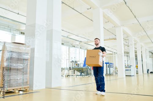 Full length portrait of handsome bearded worker wearing overall carrying cardboard box while walking along packaging department of dairy factory