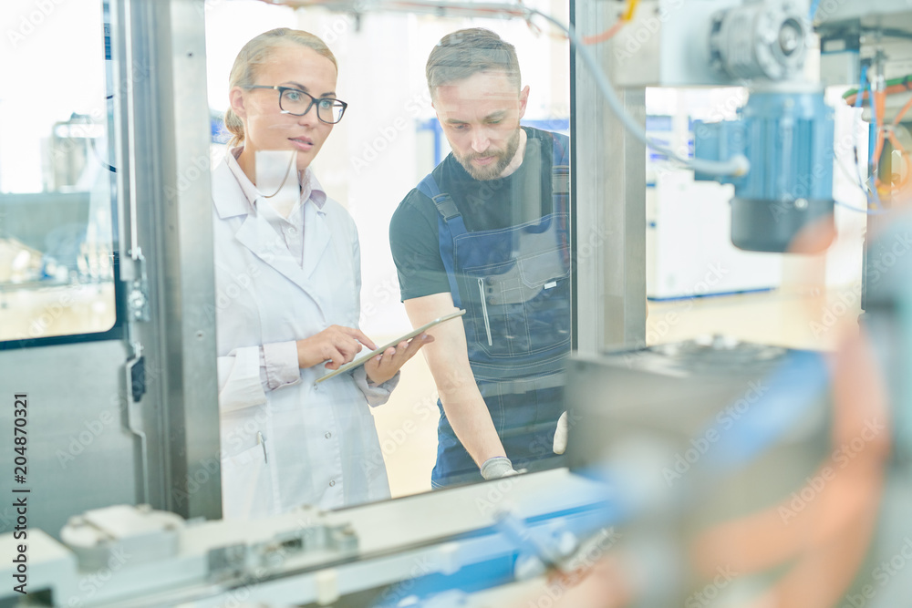 Usual working day at dairy factory: pretty blonde machine operator wearing white coat using digital tablet while bearded technician adjusting equipment