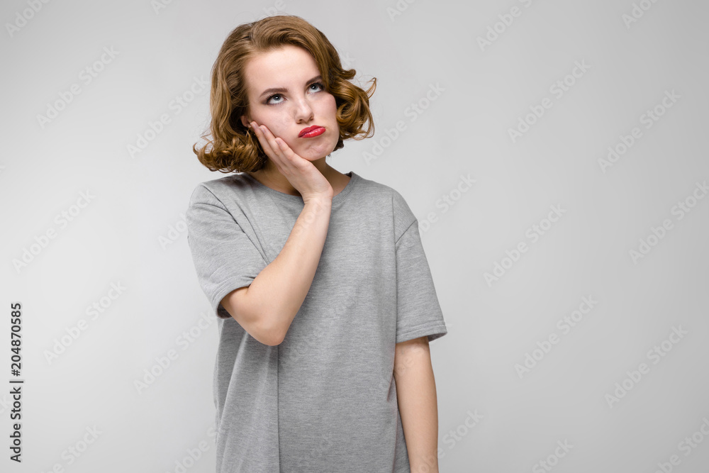 Charming young girl in a gray T-shirt on a gray background. The girl holds her hand to her cheek Charming young girl in a gray T-shirt on a gray background. The girl holds her hand to her cheek