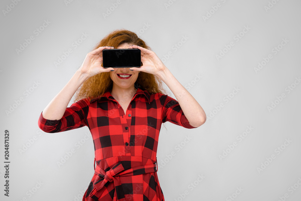 Young red-haired girl in a red checkered shirt. A young girl covered her eyes with a phone Young red-haired girl in a red checkered shirt. A young girl covered her eyes with a phone