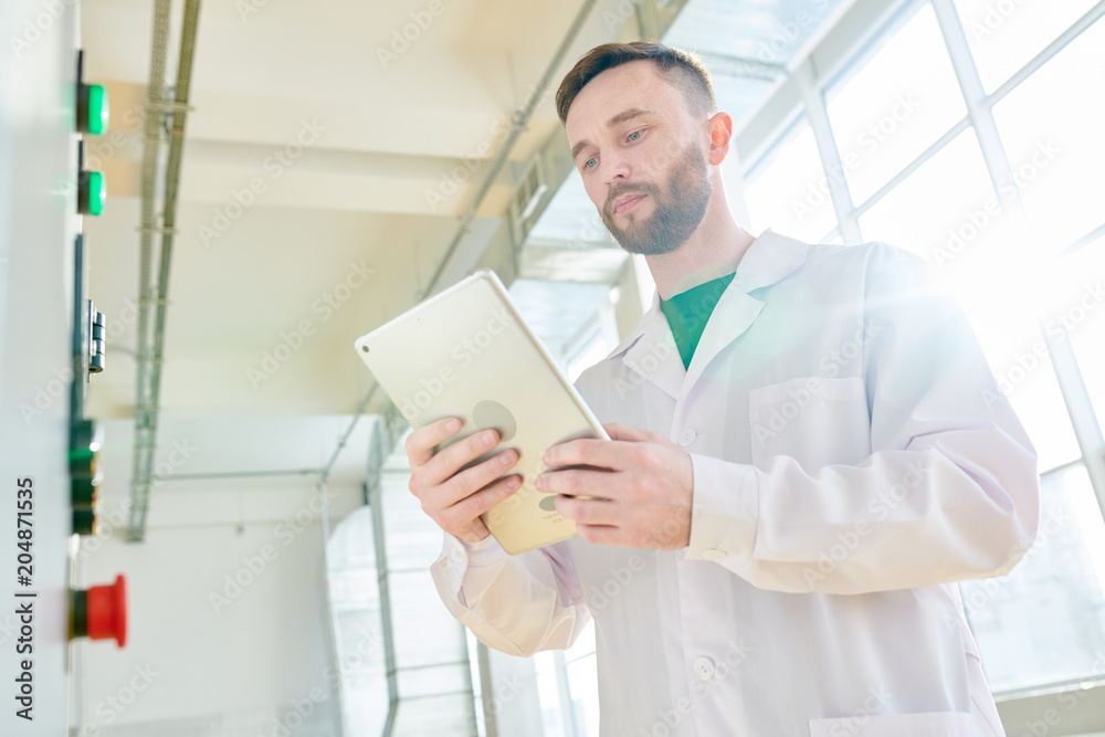 Low angle view of concentrated technologist taking necessary notes with help of digital tablet while standing at production department of dairy factory